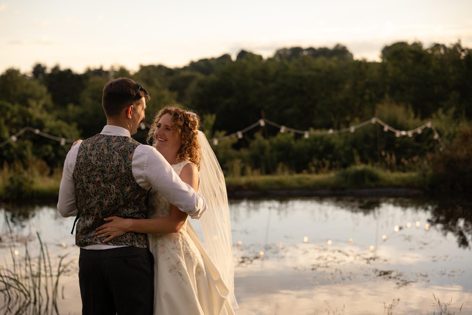 Bride and groom laughing together beside a pond at golden hour, festoon lights strung above the water